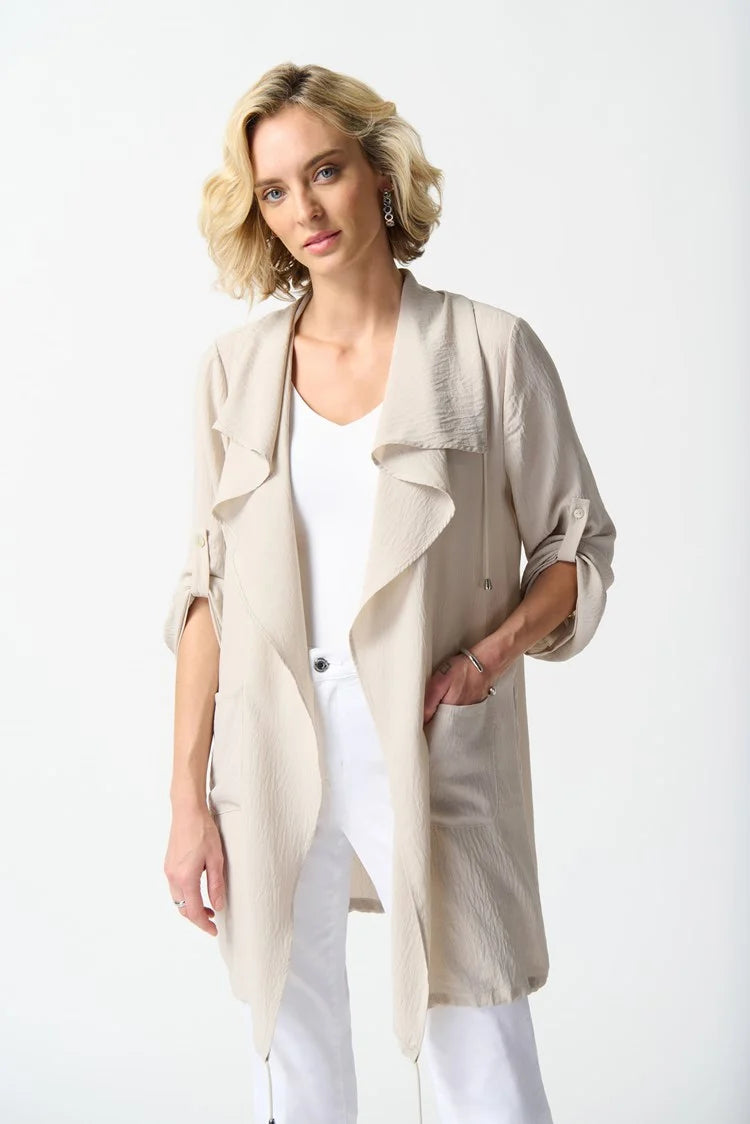 A person stands against a plain white background wearing the Joseph Ribkoff Gauze Boxy Cover Up in Moonstone with a shawl collar over a white top, white pants, and white heeled sandals. The individual has short blonde hair styled in loose waves and poses with their head turned slightly to the right.