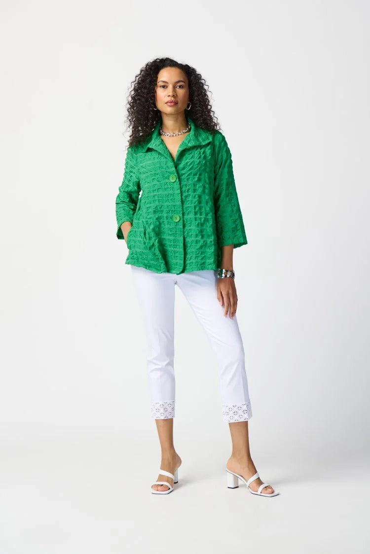 A woman with curly hair is wearing a bright green textured jacket with a button closure, white Joseph Ribkoff Millennium Crop Pull-on Pants (Style 241102) that feature lace trim at the hem, and white open-toe sandals. She is standing against a plain white background with one hand in her pocket.