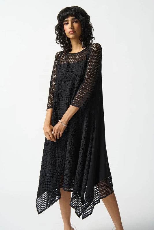 A person with wavy dark hair is wearing the Joseph Ribkoff Silky knit and Seersucker Handkerchief Dress in Black, featuring a unique geometric net-like pattern. The dress includes three-quarter sleeves and a boat neckline, enhancing its asymmetrical look. The individual stands against a plain white background with a confident expression.