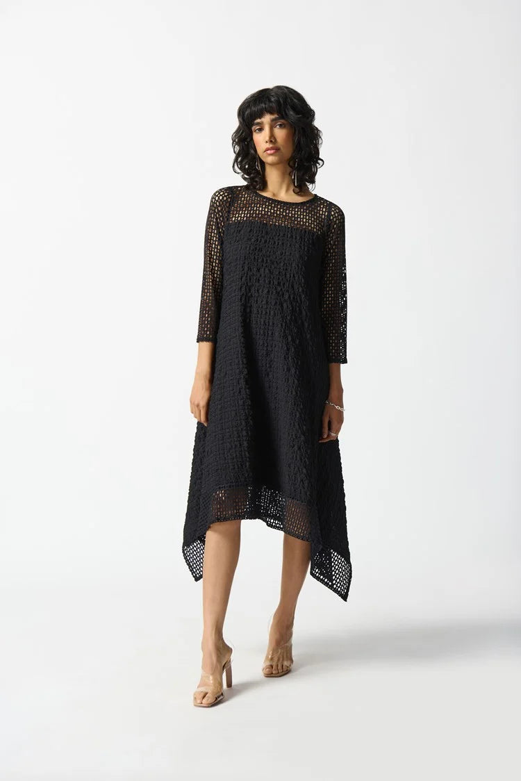 A person with wavy dark hair is wearing the Joseph Ribkoff Silky knit and Seersucker Handkerchief Dress in Black, featuring a unique geometric net-like pattern. The dress includes three-quarter sleeves and a boat neckline, enhancing its asymmetrical look. The individual stands against a plain white background with a confident expression.