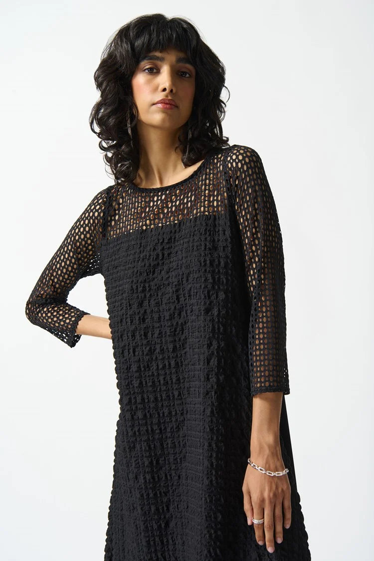 A person with wavy dark hair is wearing the Joseph Ribkoff Silky knit and Seersucker Handkerchief Dress in Black, featuring a unique geometric net-like pattern. The dress includes three-quarter sleeves and a boat neckline, enhancing its asymmetrical look. The individual stands against a plain white background with a confident expression.