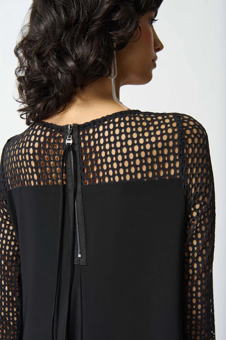 A person with wavy dark hair is wearing the Joseph Ribkoff Silky knit and Seersucker Handkerchief Dress in Black, featuring a unique geometric net-like pattern. The dress includes three-quarter sleeves and a boat neckline, enhancing its asymmetrical look. The individual stands against a plain white background with a confident expression.