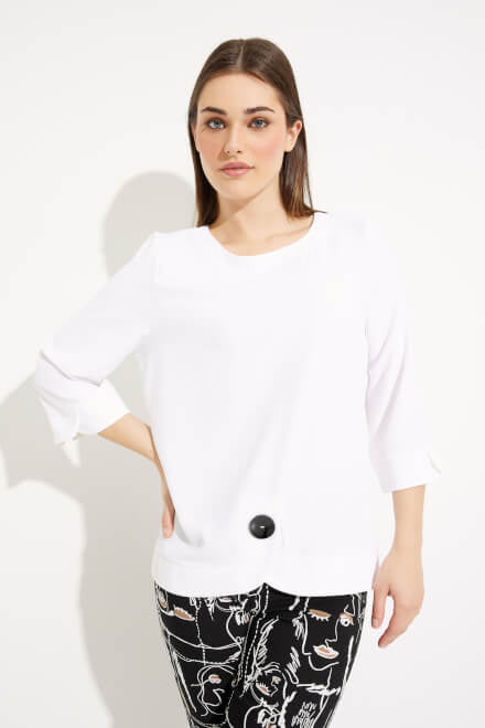 A woman with long brown hair stands against a white background, wearing a white Joseph Ribkoff Button Detail Top Style 233206 with three-quarter sleeves. She also has on black pants featuring abstract white line drawings of faces.