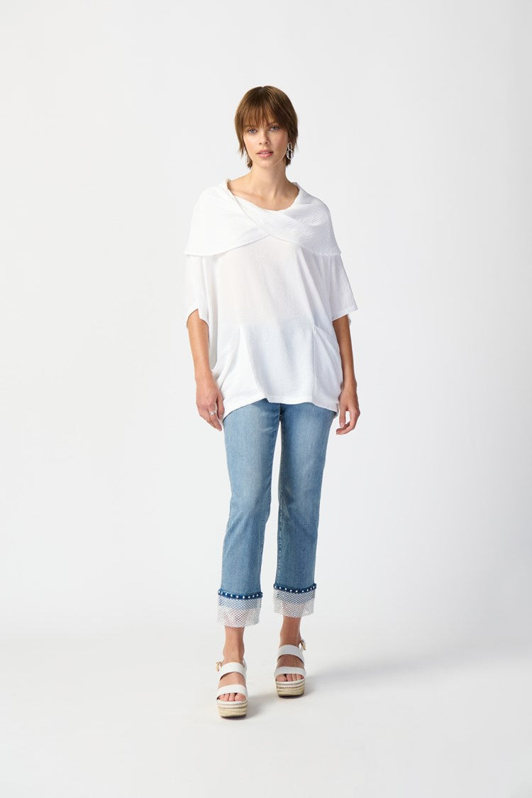 Against a plain white background, a person with short hair is seen wearing the loose-fitting Joseph Ribkoff Gauze Cowl Neck Tunic in White (242043), paired with light blue cropped jeans featuring frayed hems, and white platform sandals. The individual gazes forward with a neutral expression and relaxed posture.