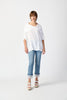 Against a plain white background, a person with short hair is seen wearing the loose-fitting Joseph Ribkoff Gauze Cowl Neck Tunic in White (242043), paired with light blue cropped jeans featuring frayed hems, and white platform sandals. The individual gazes forward with a neutral expression and relaxed posture.