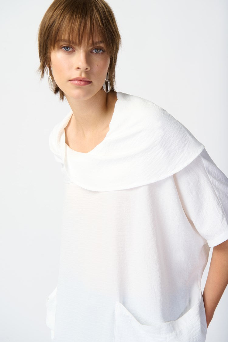 Against a plain white background, a person with short hair is seen wearing the loose-fitting Joseph Ribkoff Gauze Cowl Neck Tunic in White (242043), paired with light blue cropped jeans featuring frayed hems, and white platform sandals. The individual gazes forward with a neutral expression and relaxed posture.