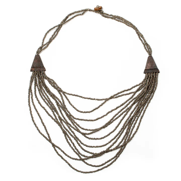 Introducing the Suzie Blue Multi Strand Wooden Side Necklace - GREY: This ethically sourced, handmade piece showcases multi-strand layers of small metallic beads, elegantly accented with two triangular brass details on either side. The varying lengths of the strands create a stunning cascading effect and it's secured with a wooden clasp at the back for easy fastening.