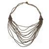 Introducing the Suzie Blue Multi Strand Wooden Side Necklace - GREY: This ethically sourced, handmade piece showcases multi-strand layers of small metallic beads, elegantly accented with two triangular brass details on either side. The varying lengths of the strands create a stunning cascading effect and it's secured with a wooden clasp at the back for easy fastening.