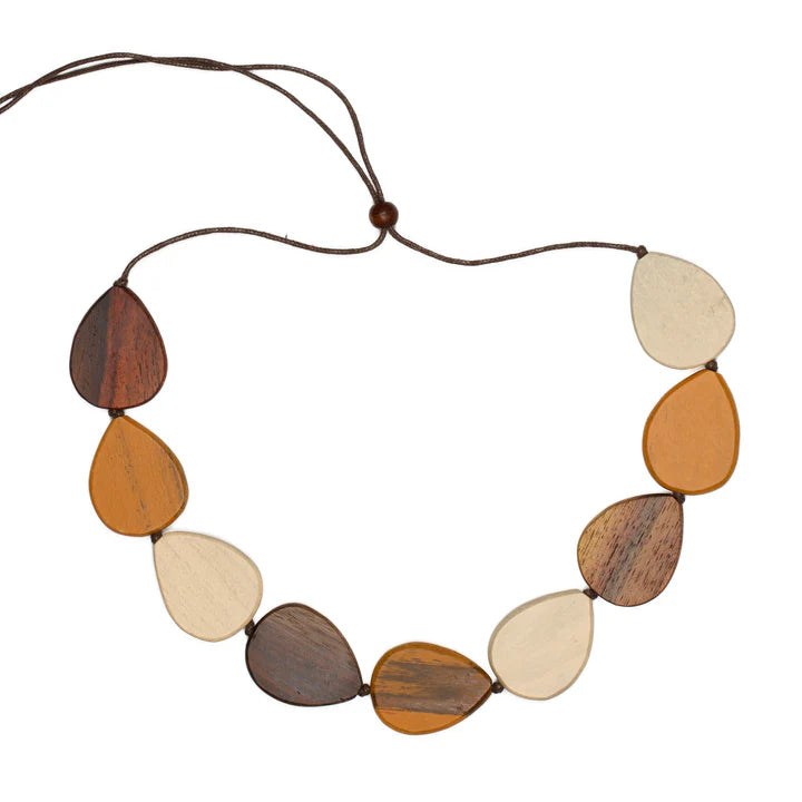 This Suzie Blue Tinted Wood Pebble Necklace showcases eight teardrop-shaped pendants in varying shades of brown, cream, and beige, all strung on a dark brown cord. Made from ethically sourced wood, each pendant's unique tint gives the piece an earthy, natural look.