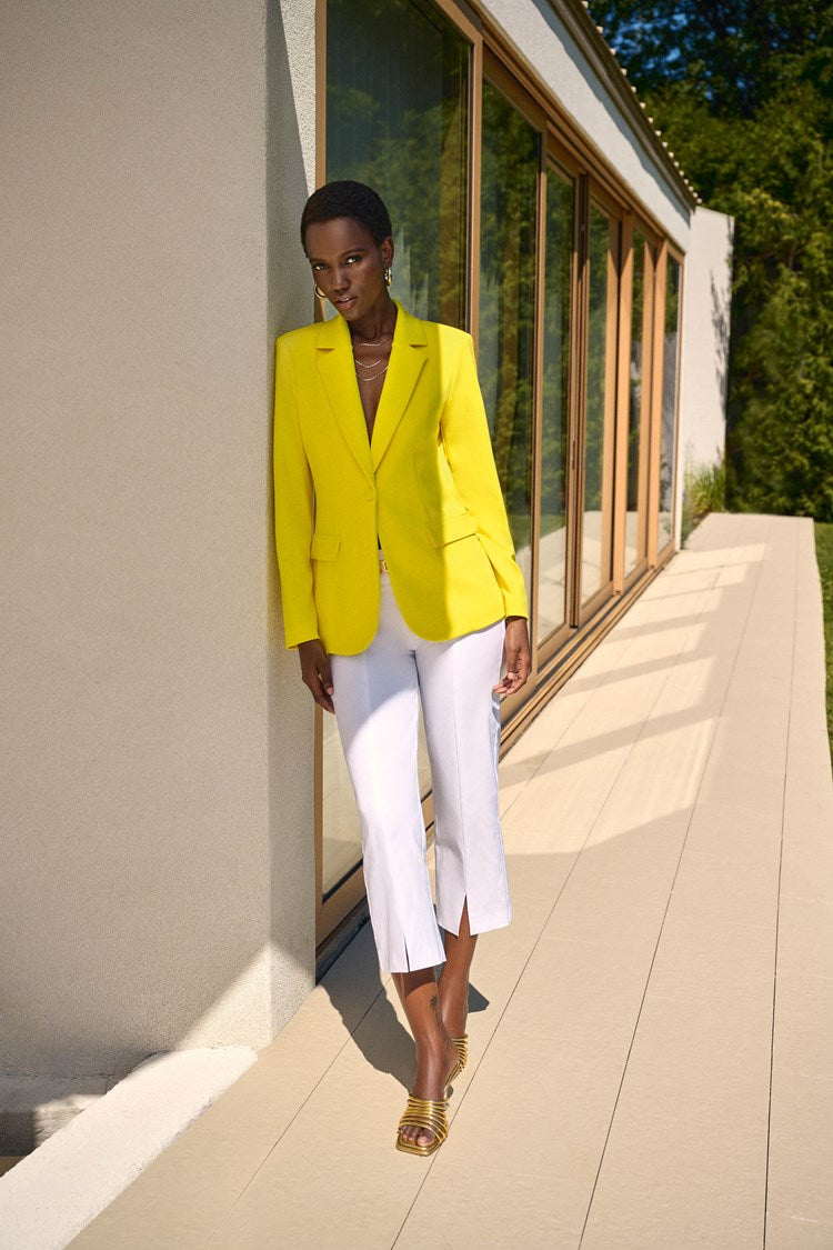 A person with short blonde hair is wearing a bright yellow blazer over a white top, Millennium Straight Crop Pants 242035 from Joseph Ribkoff, and matching yellow shoes. They accessorize with gold earrings and a necklace and have a calm expression while posing against a plain white background.