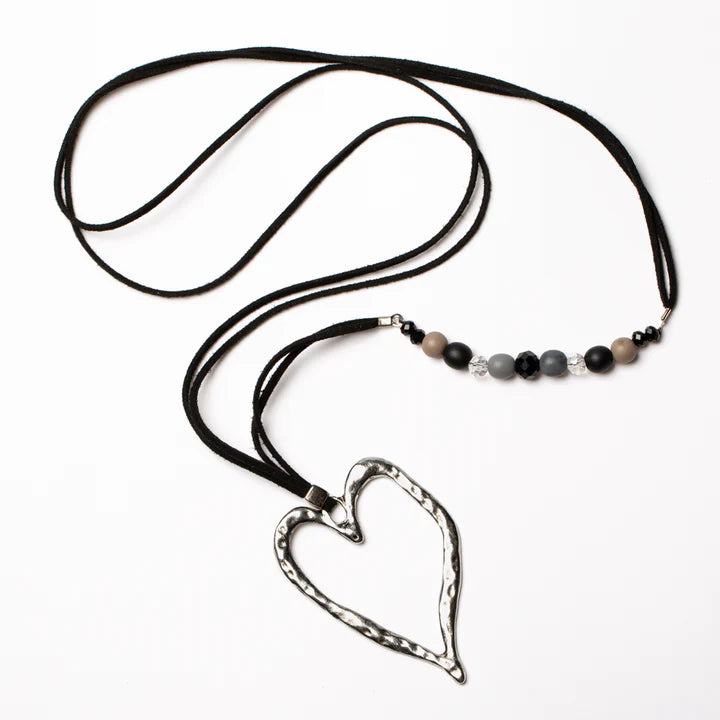 The Long Open Heart Necklace with Offset Beads - BLACK by Suzie Blue is a stunning piece featuring a large, hammered silver heart pendant as its focal point. This handmade necklace incorporates beads in varying shades of black, gray, and beige, set off beautifully against the long black leather cord. Crafted using ethically sourced materials, the design is simple yet elegant.