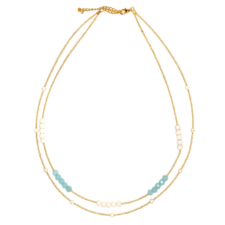 The Double Strand Necklace With Crystal Beads by Suzie Blue is a handmade gold chain piece featuring small white and blue beads arranged in clusters. This delicate necklace includes an adjustable clasp at the back for customizable length, enhancing its simple yet elegant design.