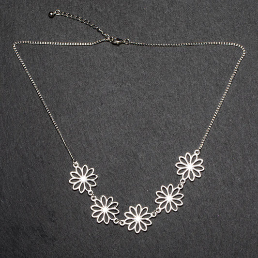 The Suzie Blue "Five Flower Necklace In Silver Plate," part of our handmade jewelry collection, showcases a delicate chain with an adjustable clasp and five daisy-shaped floral pendants beautifully linked together in the center. Set against a plain, dark, textured surface, the intricate design of this ethically sourced piece truly stands out.