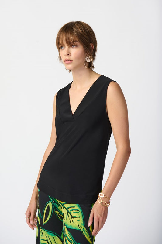 A person with short brown hair is wearing a Silky Knit Sleeveless Top 241239 by Joseph Ribkoff in black, along with large hoop earrings. They are also sporting a bracelet and a skirt featuring a green leaf pattern. The background is plain white.