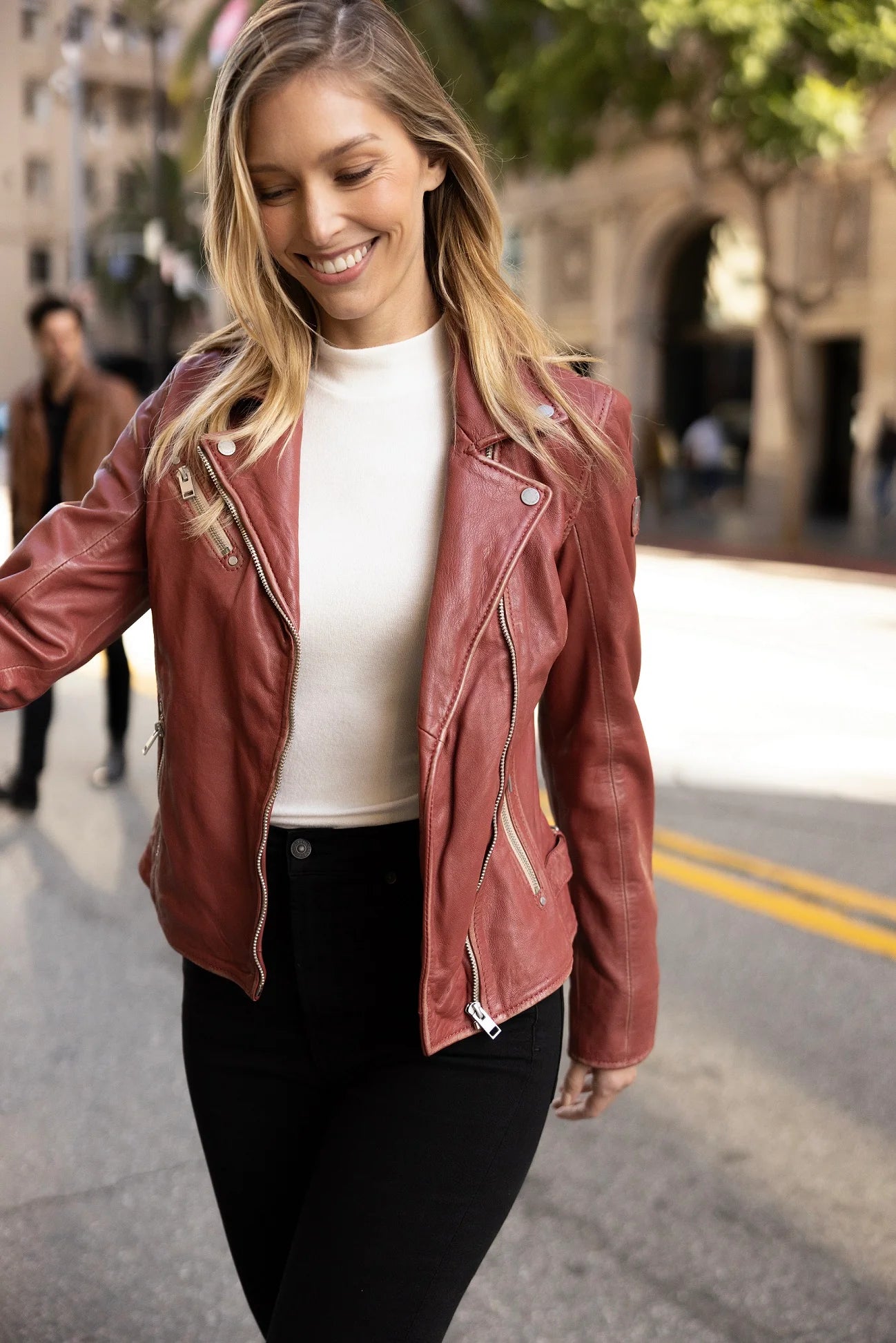 A smiling woman with long blonde hair walks down a city street, epitomizing true ladies' style. She is wearing a Sofia RF Leather Jacket in Astro Dust from MAURITIUS LEATHER over a white top and black pants. The street is lined with trees and buildings, with another person visible in the background.