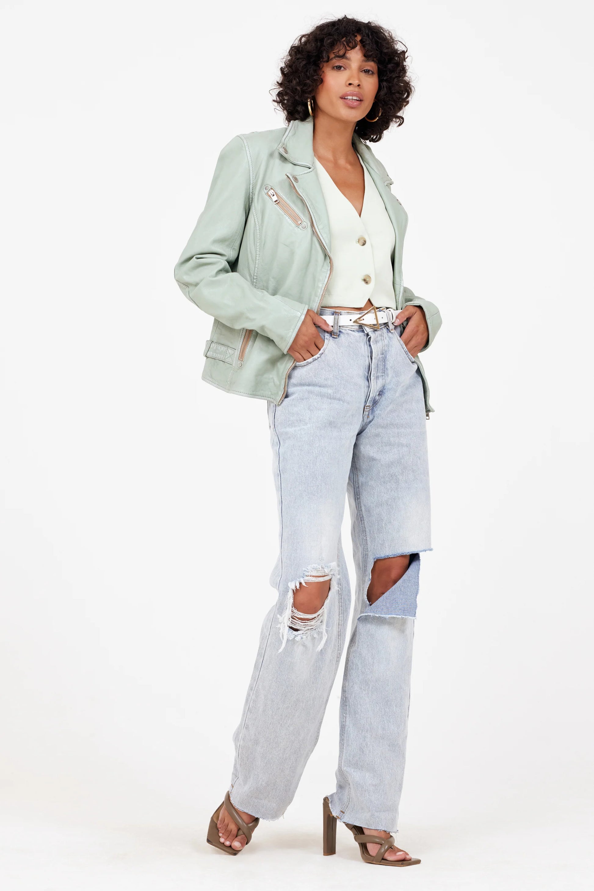 A person with curly hair stands confidently against a white background. They wear the Sofia RF Leather Jacket in Frosty Green from MAURITIUS LEATHER over a white top, paired with distressed light blue jeans and heeled sandals. Their hands are placed in their jacket pockets as they look straight ahead.