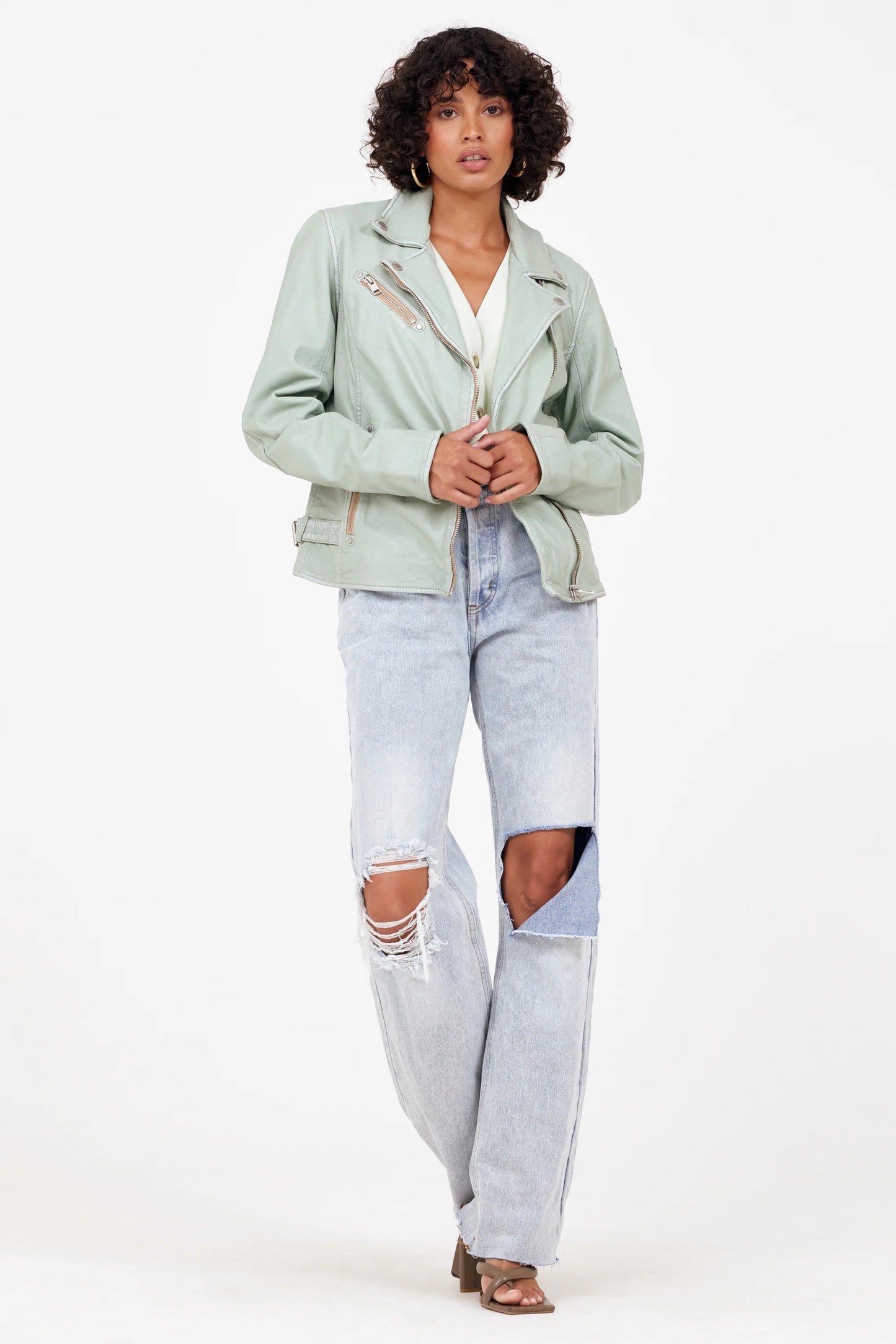 A person with curly hair stands confidently against a white background. They wear the Sofia RF Leather Jacket in Frosty Green from MAURITIUS LEATHER over a white top, paired with distressed light blue jeans and heeled sandals. Their hands are placed in their jacket pockets as they look straight ahead.