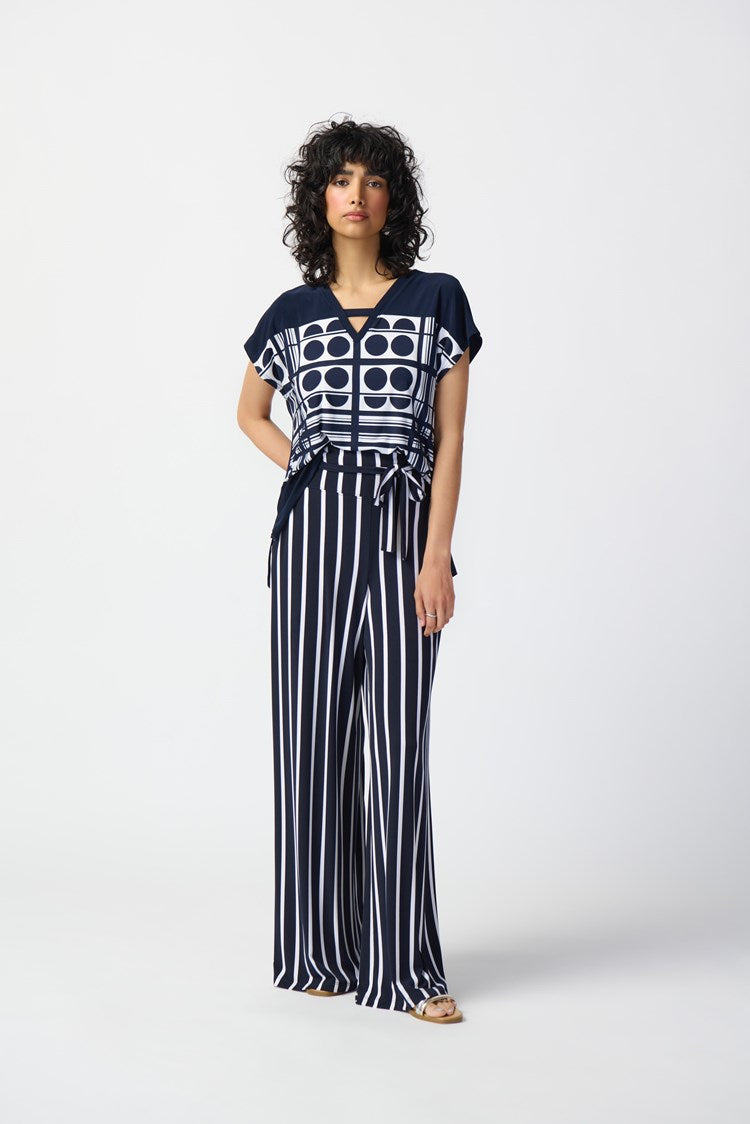 A person stands against a plain white background wearing a stylish outfit from Joseph Ribkoff. The top is the Geometric Print Silky Knit V-Neck Top 241117, featuring short sleeves, predominantly midnight blue with vanilla circular graphic prints. The high-waisted pants have vertical navy and white stripes, completed by a tied waist belt.