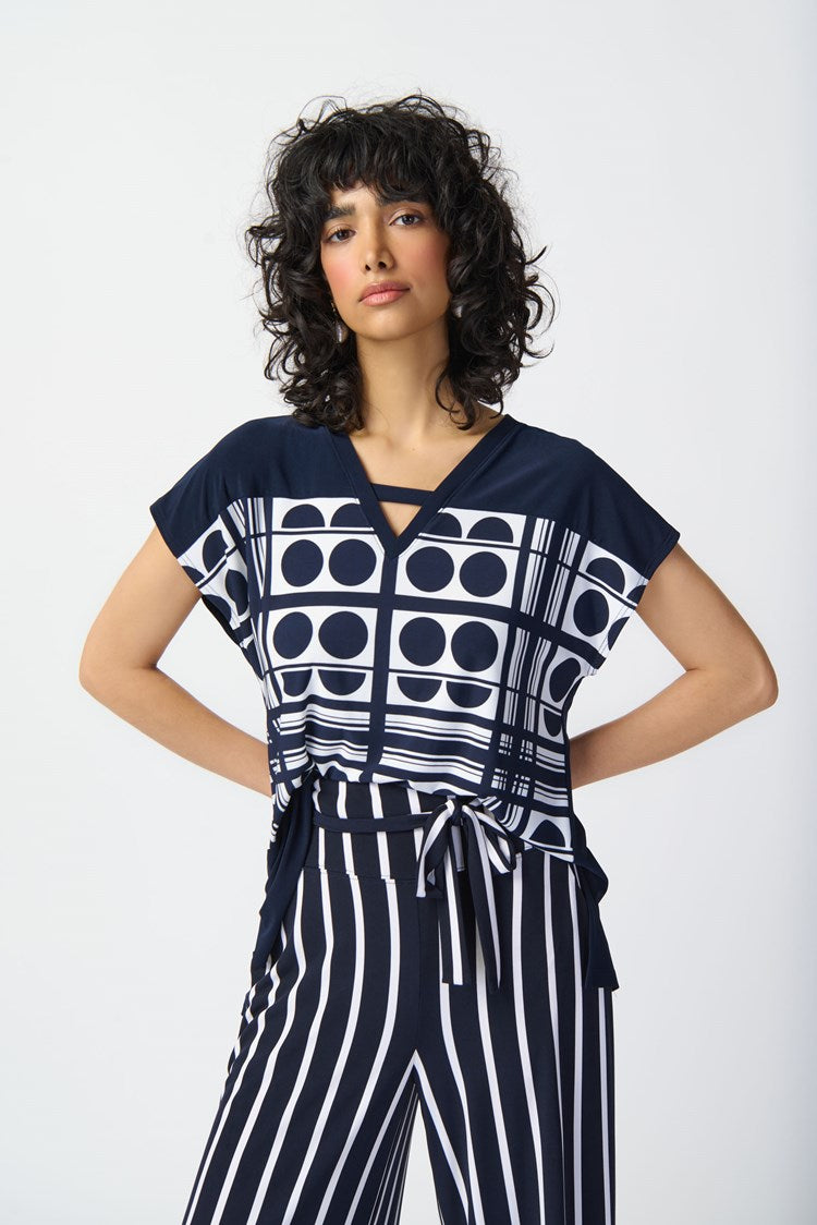 A person stands against a plain white background wearing a stylish outfit from Joseph Ribkoff. The top is the Geometric Print Silky Knit V-Neck Top 241117, featuring short sleeves, predominantly midnight blue with vanilla circular graphic prints. The high-waisted pants have vertical navy and white stripes, completed by a tied waist belt.