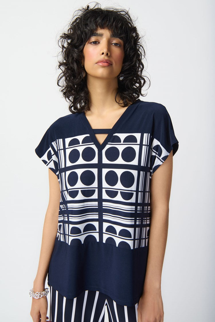 A person stands against a plain white background wearing a stylish outfit from Joseph Ribkoff. The top is the Geometric Print Silky Knit V-Neck Top 241117, featuring short sleeves, predominantly midnight blue with vanilla circular graphic prints. The high-waisted pants have vertical navy and white stripes, completed by a tied waist belt.