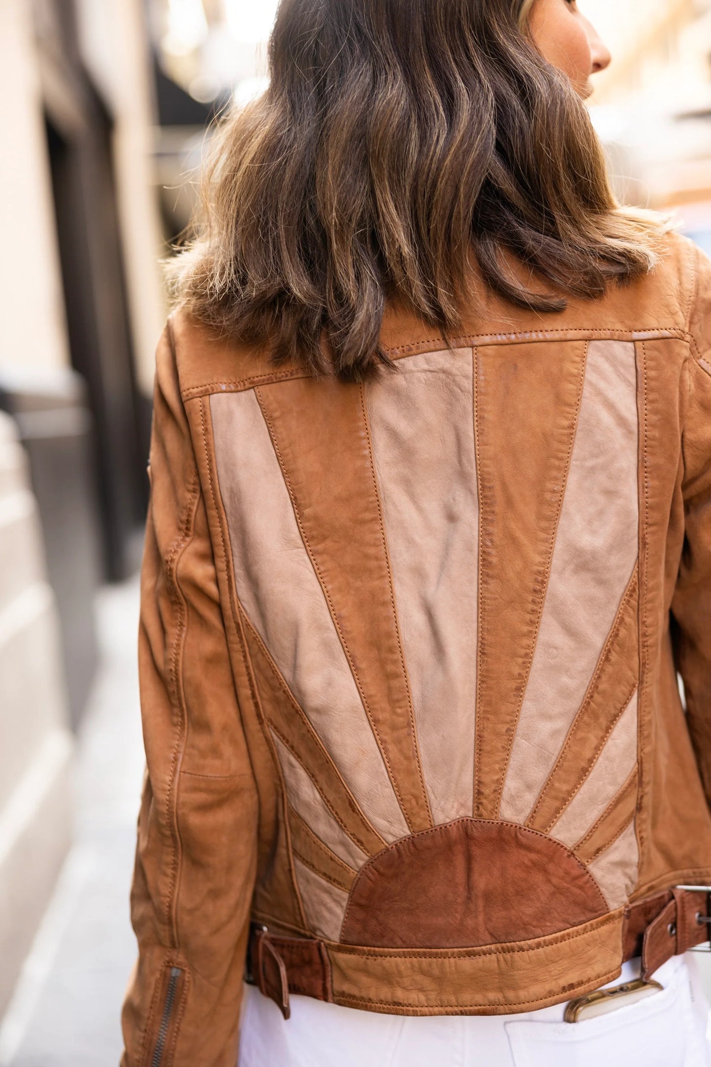 A person with shoulder-length brown hair faces away from the camera, wearing the Sunny RF Leather Jacket in cognac by MAURITIUS LEATHER, which showcases varying shades of tan and brown with a sunset design featuring rays emanating from a circular center at the bottom.