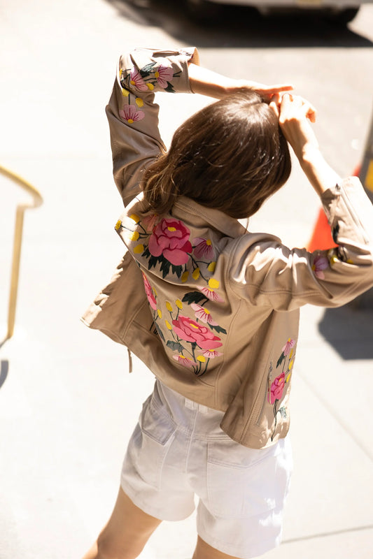 A person with long hair is seen from behind, adjusting their hair. They are wearing a light beige Peonie RF Leather Jacket by MAURITIUS LEATHER, adorned with colorful floral embroidery on the back and sleeves, paired with white shorts. The scene is bright and sunny, suggesting it is taken outdoors.