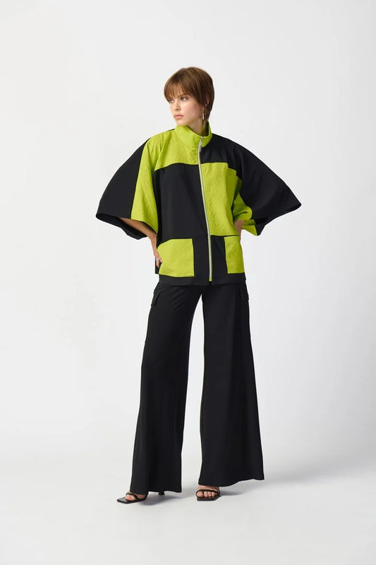 A person with short hair wears a stylish, oversized Joseph Ribkoff Silky Knit And Jacquard Boxy Jacket 241198 in Black/Key Lime, featuring three-quarter dolman sleeves and a funnel collar. The outfit is paired with wide-leg black pants and black open-toe heels against a plain white backdrop.