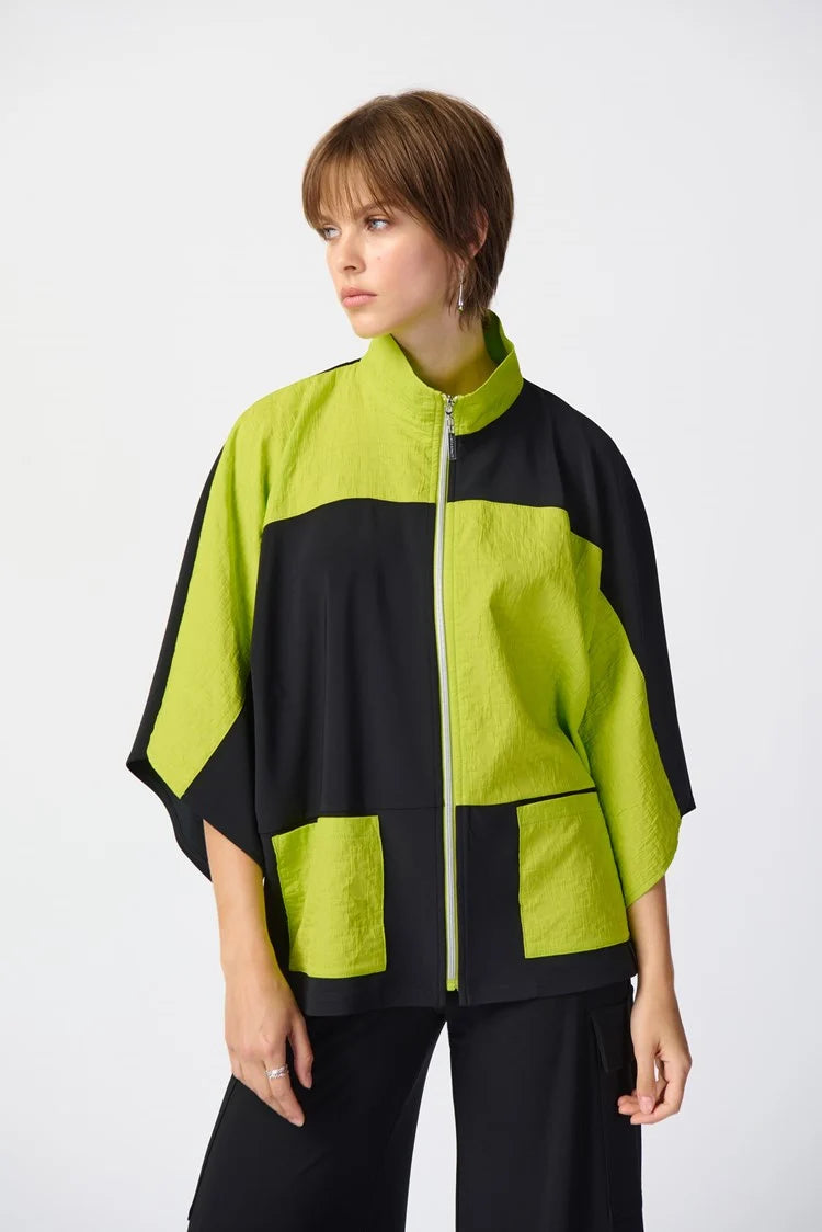 A person with short hair wears a stylish, oversized Joseph Ribkoff Silky Knit And Jacquard Boxy Jacket 241198 in Black/Key Lime, featuring three-quarter dolman sleeves and a funnel collar. The outfit is paired with wide-leg black pants and black open-toe heels against a plain white backdrop.