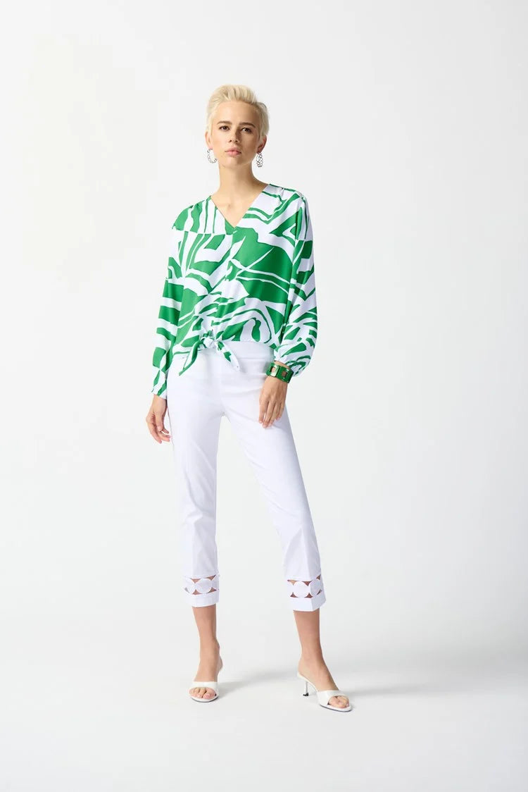 A person with short blonde hair is wearing a Joseph Ribkoff Woven Abstract Print Front Tie Top 242121 in Green/Vanilla, paired with white capri pants featuring a decorative cutout at the cuffs. They are standing with one foot slightly forward and are wearing white open-toe heels. The background is a plain, light color.