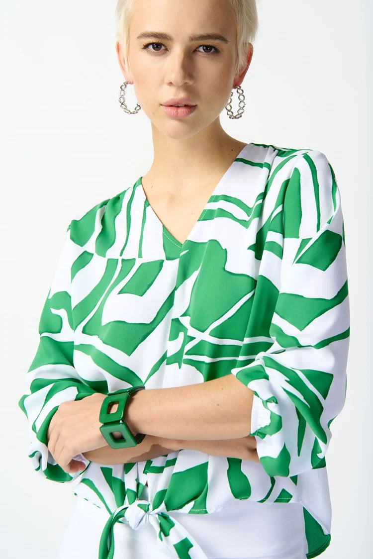 A person with short blonde hair is wearing a Joseph Ribkoff Woven Abstract Print Front Tie Top 242121 in Green/Vanilla, paired with white capri pants featuring a decorative cutout at the cuffs. They are standing with one foot slightly forward and are wearing white open-toe heels. The background is a plain, light color.