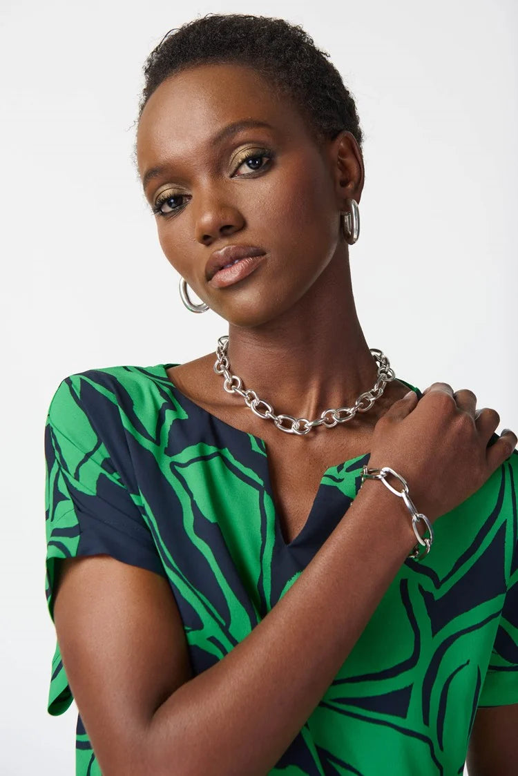 A woman with short hair is wearing the "Floral Print Georgette Boxy Top 241059 - Joseph Ribkoff - Midnight Blue/Green," a matching green and navy blue floral print top and pants set crafted from georgette fabric by Joseph Ribkoff. She is accessorized with a silver necklace and bracelets. She stands against a plain white background, looking at the camera with a neutral expression.