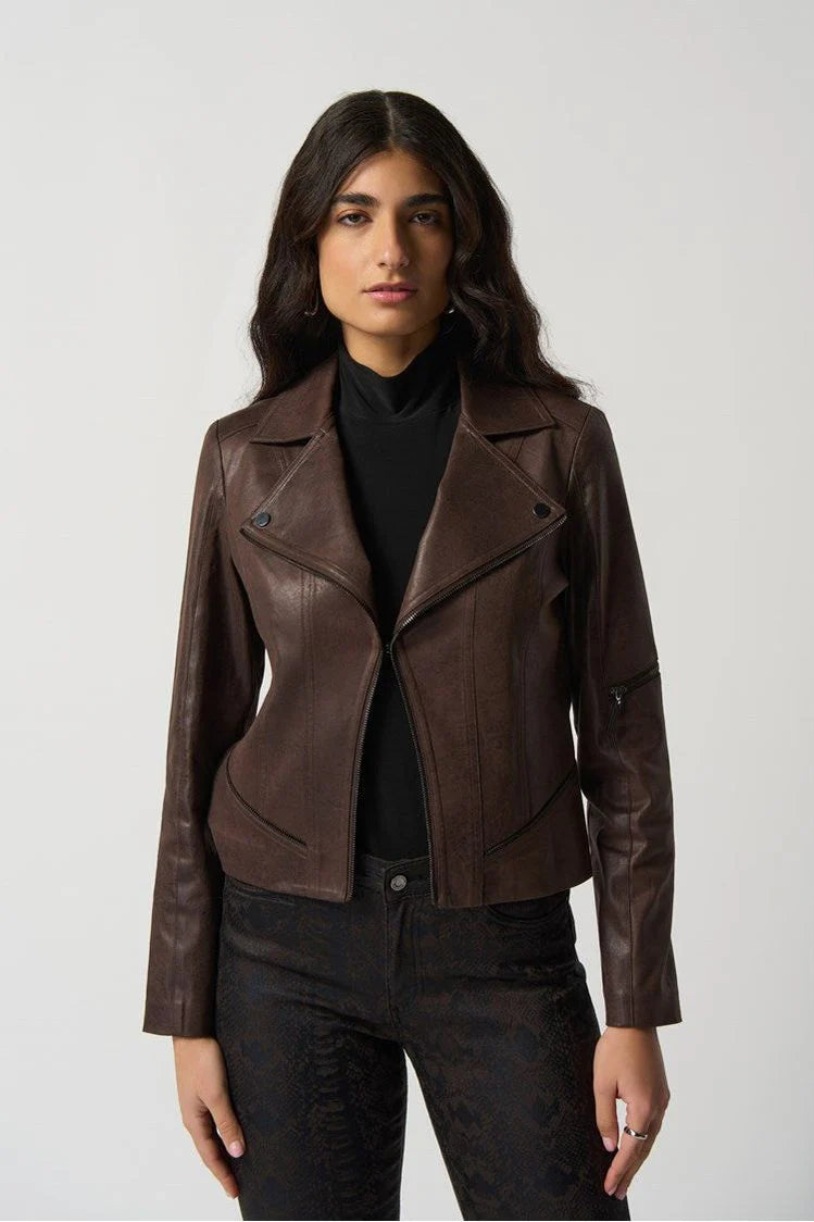 A woman with long, wavy dark hair wears the Joseph Ribkoff Pleather Moto Jacket - Style 233969 over a black turtleneck. She stands with a confident expression and her hands down by her sides, against a plain light-colored background.