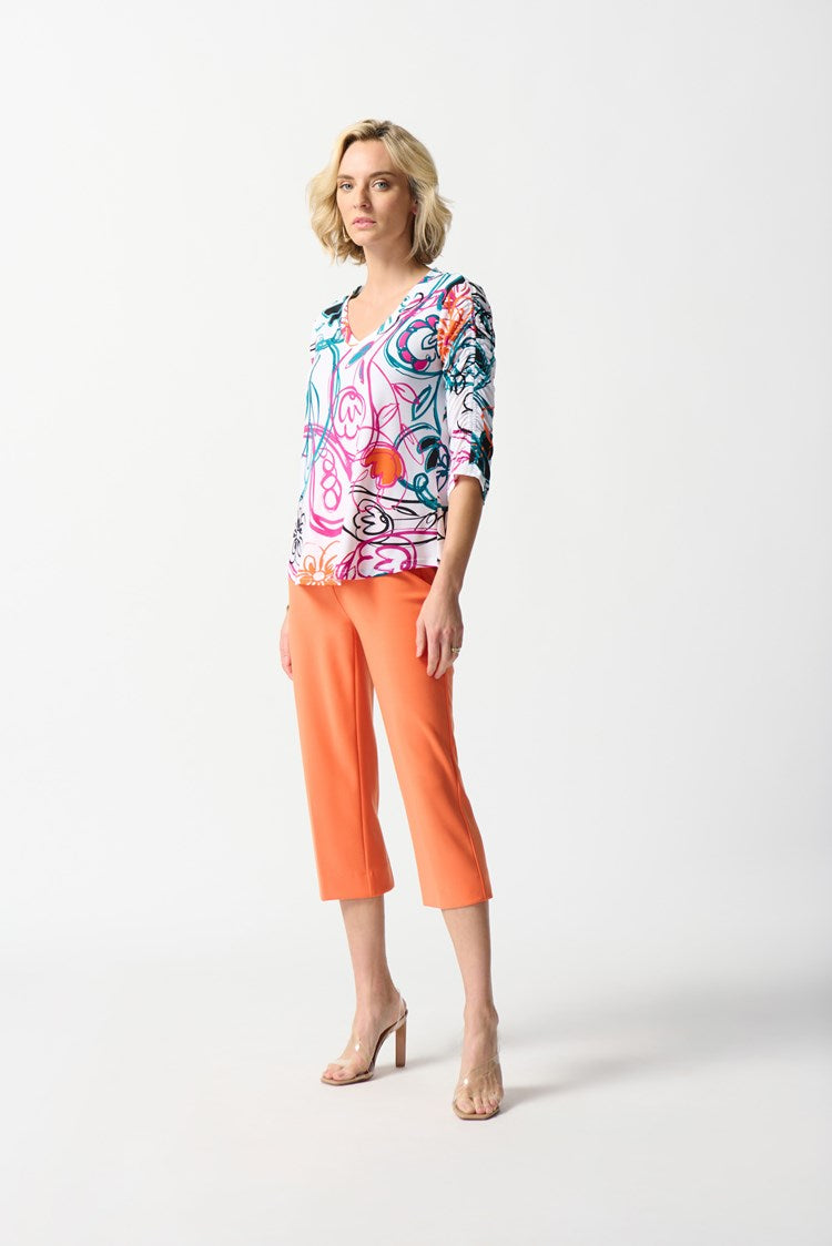 A woman with shoulder-length blonde hair stands against a white background, wearing the Joseph Ribkoff Silky Knit Paisley Print Top in Vanilla/Multi. She pairs it with bright orange cropped pants and completes her look with nude high-heeled shoes.