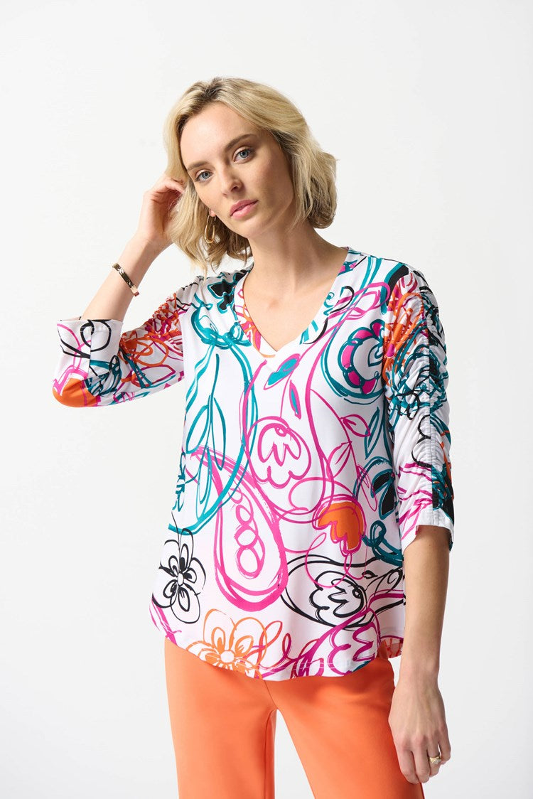 A woman with shoulder-length blonde hair stands against a white background, wearing the Joseph Ribkoff Silky Knit Paisley Print Top in Vanilla/Multi. She pairs it with bright orange cropped pants and completes her look with nude high-heeled shoes.