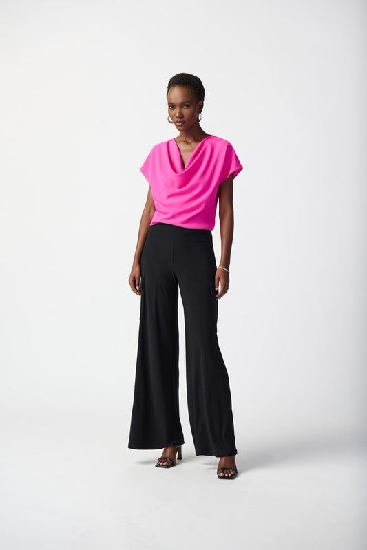 A woman stands against a plain background wearing the Joseph Ribkoff Woven Cowl Neck Top 241099 in vibrant ultra pink, paired with wide-legged black pants. She has short hair and is sporting black high-heeled sandals. Smiling slightly, she places one hand on her hip.