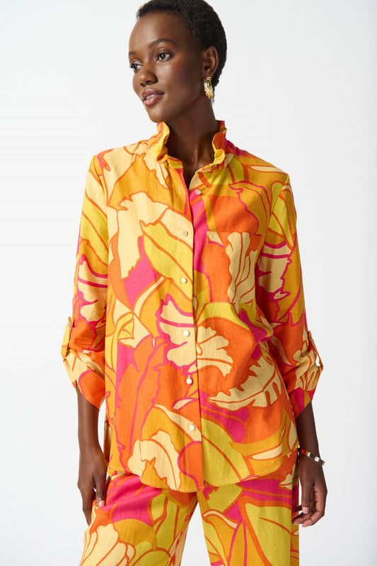 A woman wearing the Joseph Ribkoff Linen Blend Floral Print Trapeze Blouse 242915 in Pink/Multi, showcasing a vibrant ensemble with bold floral patterns in yellow, orange, and pink. She has short hair and accessorizes with gold earrings and a bracelet. The blouse features a ruffled neckline, three-quarter sleeves, and a graceful trapeze silhouette as she poses confidently.