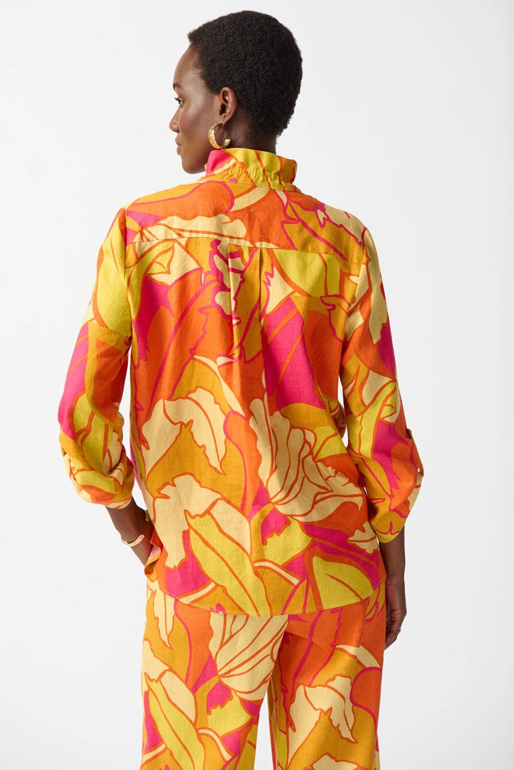 A woman wearing the Joseph Ribkoff Linen Blend Floral Print Trapeze Blouse 242915 in Pink/Multi, showcasing a vibrant ensemble with bold floral patterns in yellow, orange, and pink. She has short hair and accessorizes with gold earrings and a bracelet. The blouse features a ruffled neckline, three-quarter sleeves, and a graceful trapeze silhouette as she poses confidently.