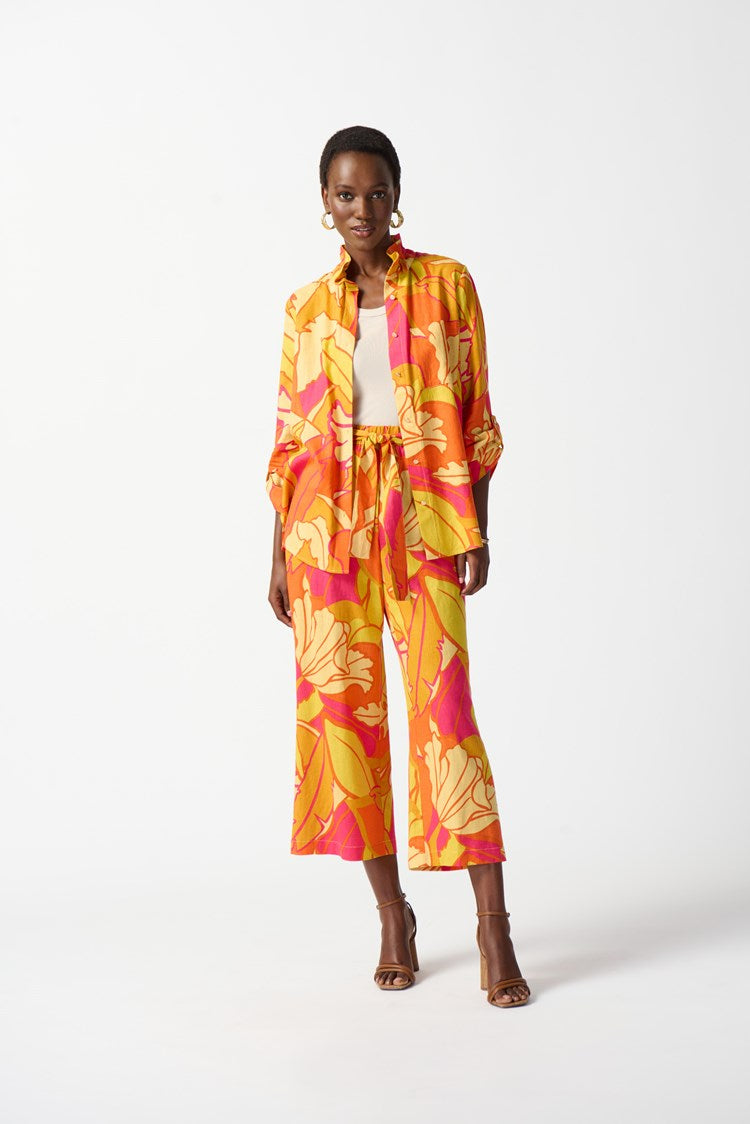 A woman wearing the Joseph Ribkoff Linen Blend Floral Print Trapeze Blouse 242915 in Pink/Multi, showcasing a vibrant ensemble with bold floral patterns in yellow, orange, and pink. She has short hair and accessorizes with gold earrings and a bracelet. The blouse features a ruffled neckline, three-quarter sleeves, and a graceful trapeze silhouette as she poses confidently.