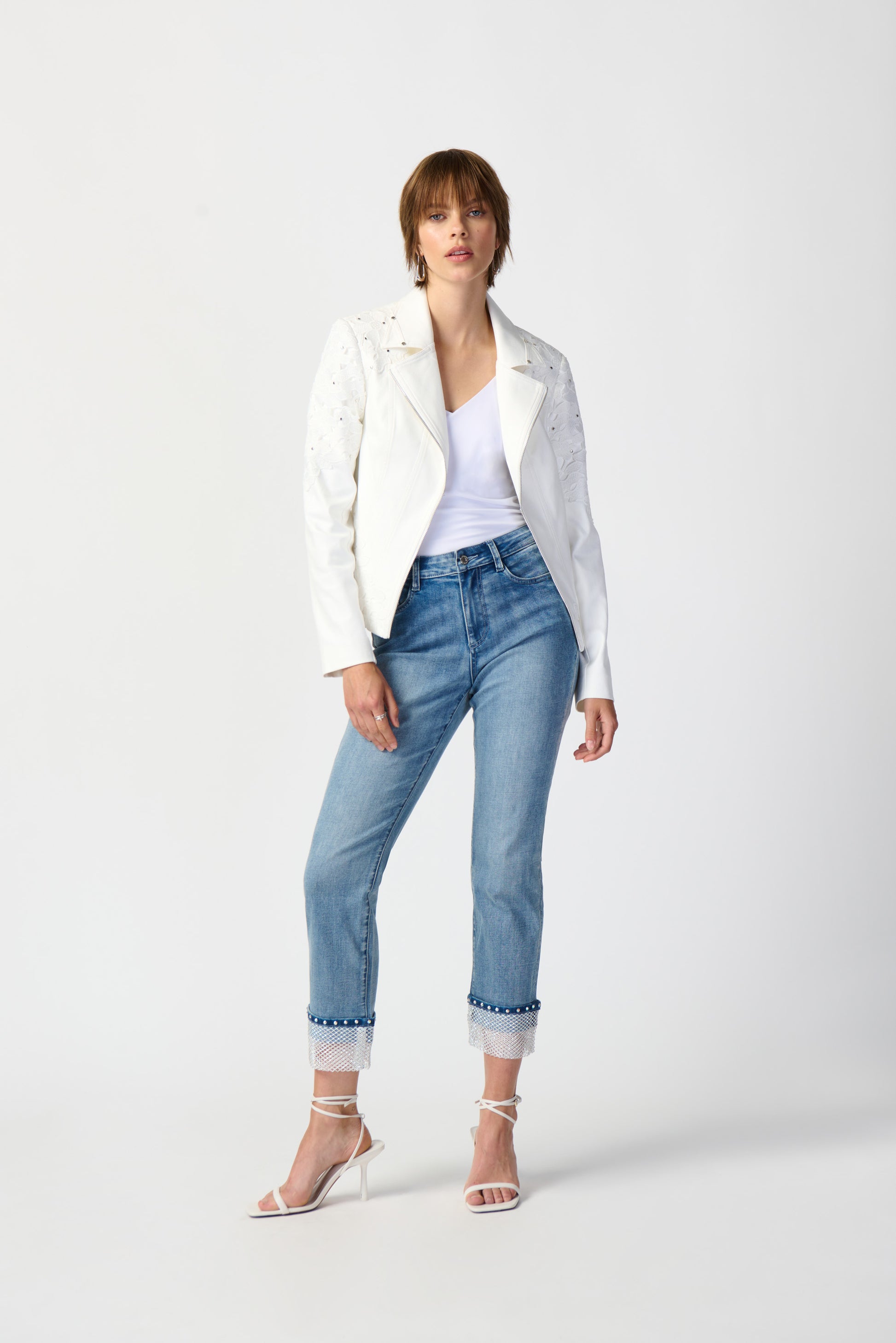 A woman stands against a plain white background wearing a Joseph Ribkoff Studded Foiled Suede Jacket with Floral Appliqué, a white top, blue cuffed jeans with embroidered hems, and white high-heeled sandals. She has short hair and is posing with one hand in her pocket.