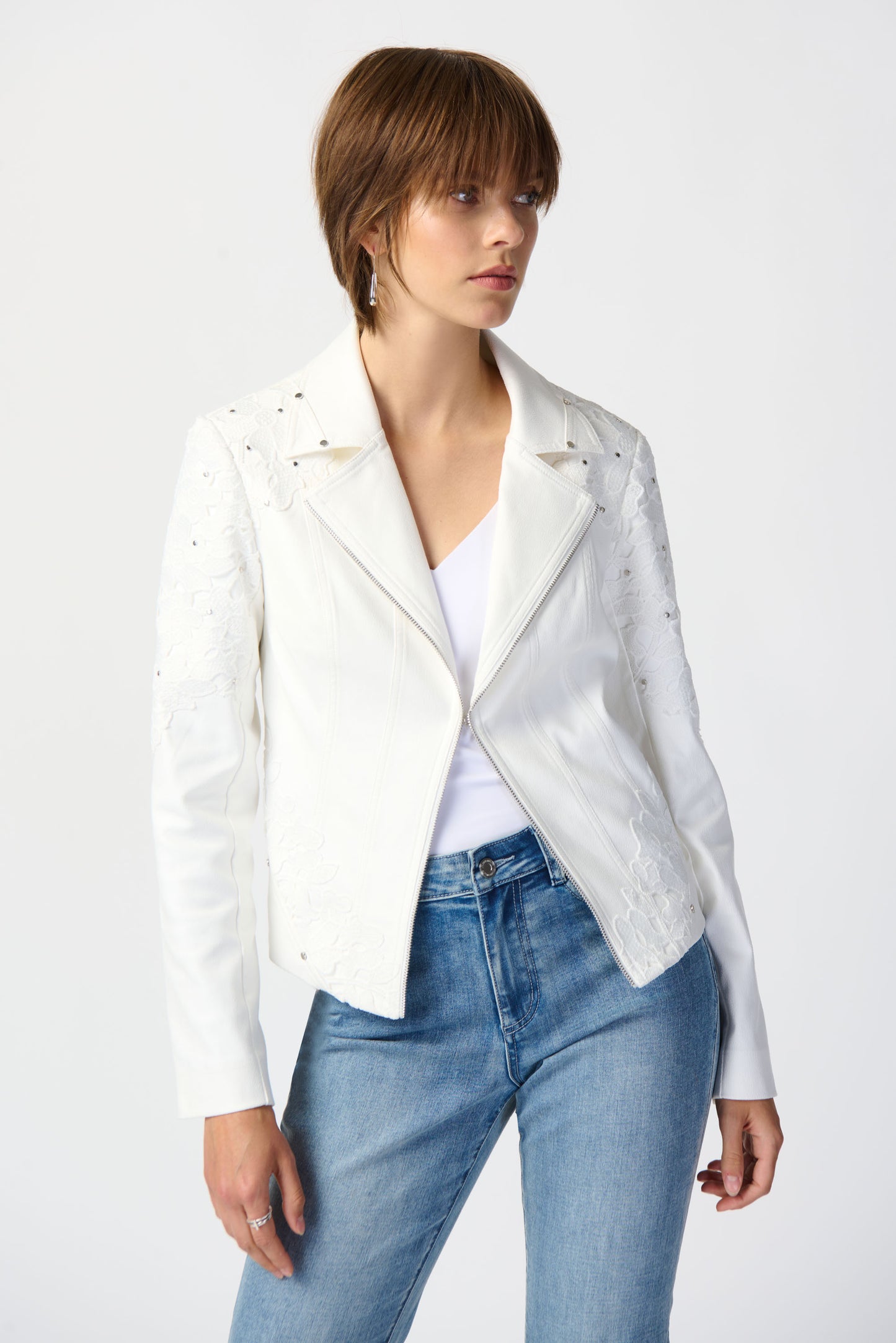 A woman stands against a plain white background wearing a Joseph Ribkoff Studded Foiled Suede Jacket with Floral Appliqué, a white top, blue cuffed jeans with embroidered hems, and white high-heeled sandals. She has short hair and is posing with one hand in her pocket.