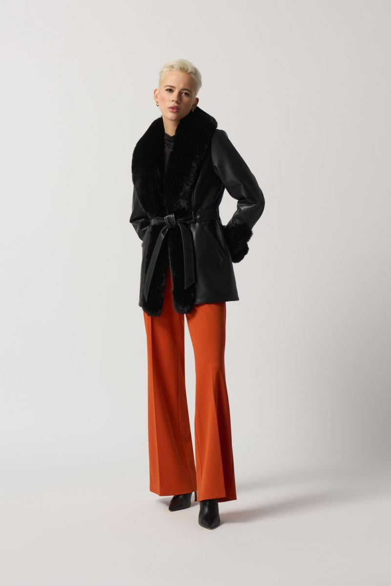 A person with short blonde hair wears the Black Leatherette Coat With Faux Fur Style 233927-Joseph Ribkoff by Joseph Ribkoff, tied at the waist with a matching belt. They have a thoughtful expression and are positioned against a plain gray background, epitomizing winter fashion at its finest.