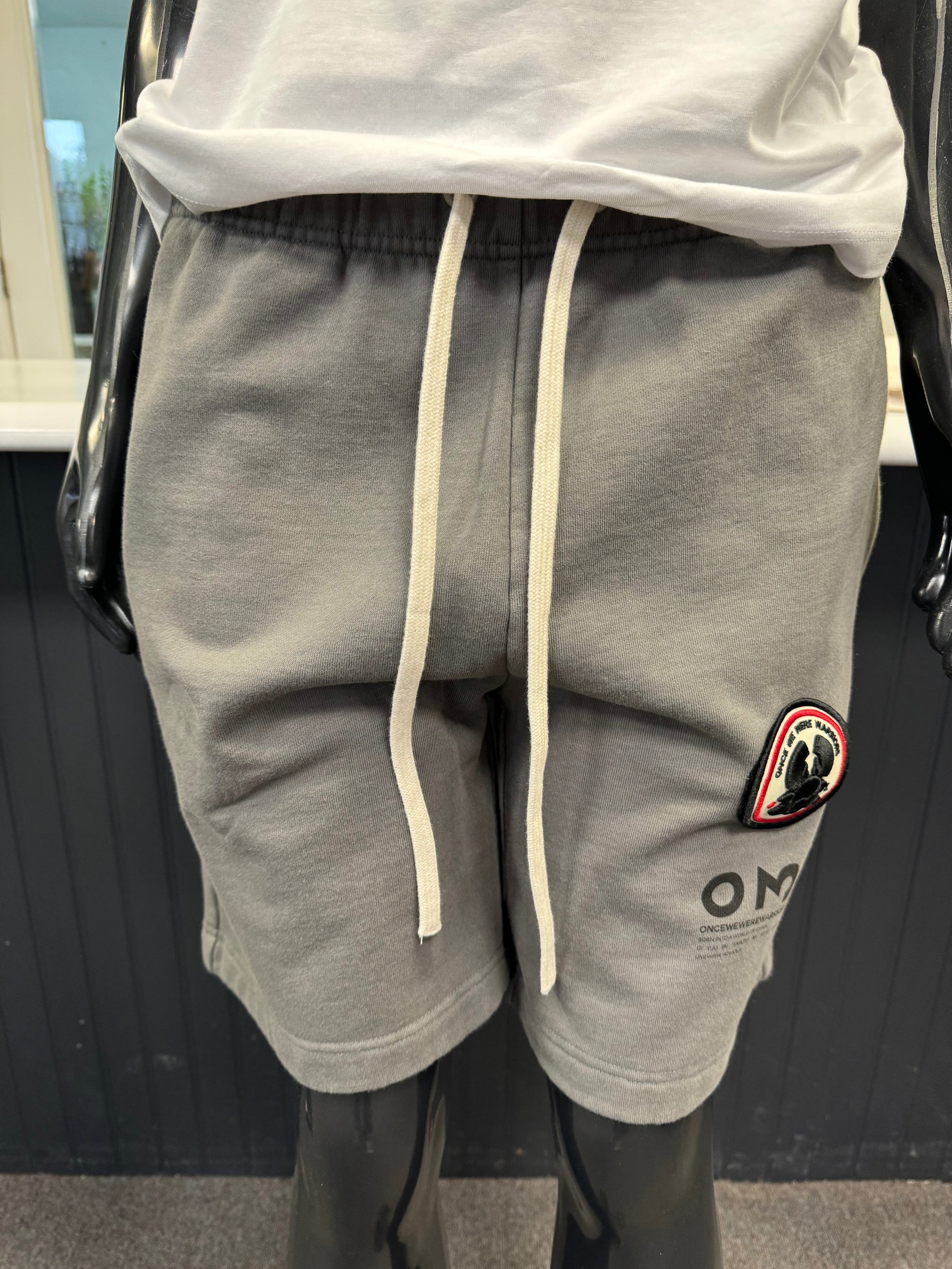 A mannequin dressed in a white t-shirt and granite grey Yabu Shorts from Once We Were Warriors is displayed. The shorts feature a small patch with a red and black design on the left leg and the letters "OMG" printed near the hem, perfectly embodying the brand's style.