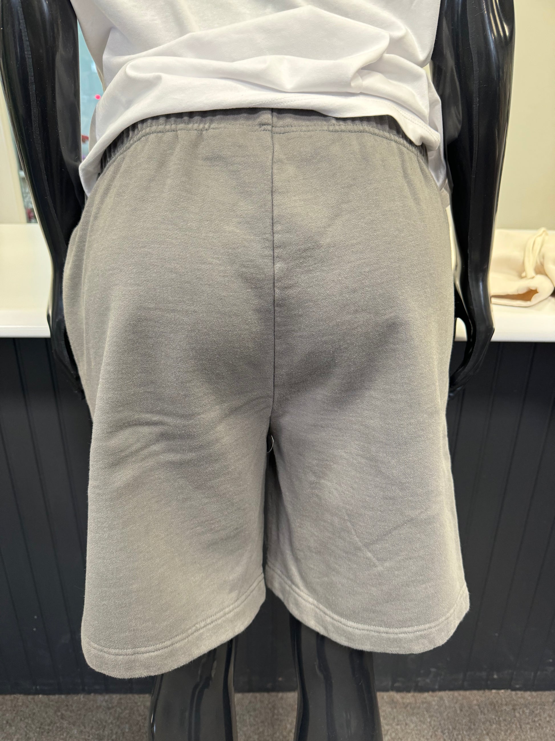 A mannequin dressed in a white t-shirt and granite grey Yabu Shorts from Once We Were Warriors is displayed. The shorts feature a small patch with a red and black design on the left leg and the letters "OMG" printed near the hem, perfectly embodying the brand's style.