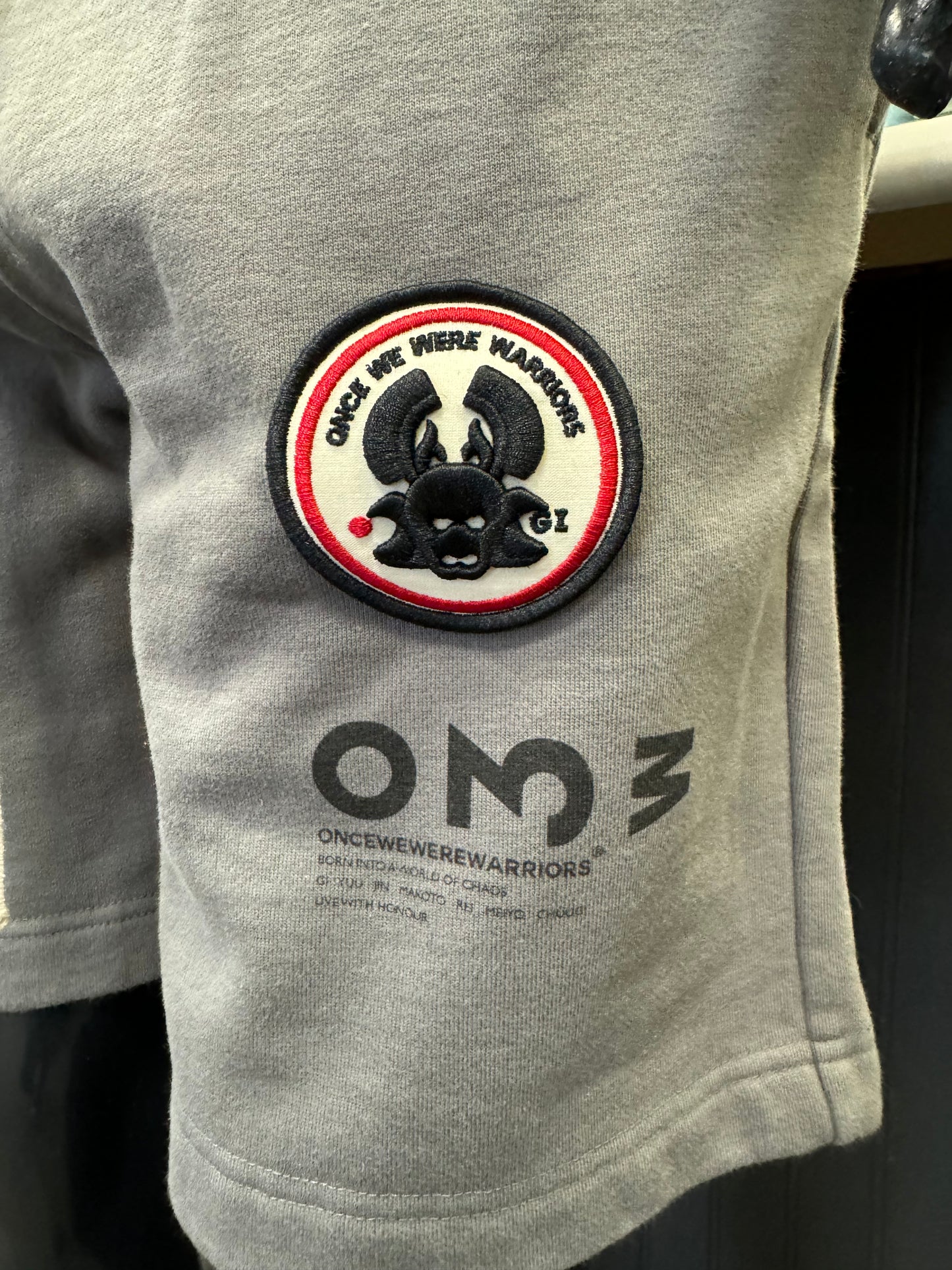 A mannequin dressed in a white t-shirt and granite grey Yabu Shorts from Once We Were Warriors is displayed. The shorts feature a small patch with a red and black design on the left leg and the letters "OMG" printed near the hem, perfectly embodying the brand's style.