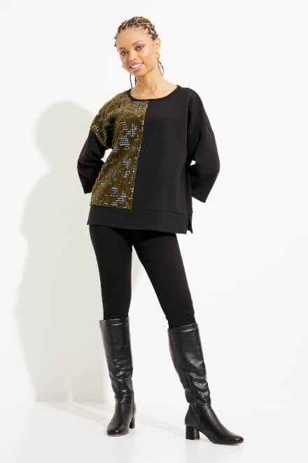 A person with braided hair stands against a plain white background wearing a stylish black long-sleeve Blocked Top Style 233267 by Joseph Ribkoff featuring a printed motif on one side, black pants, and knee-high boots with heels. They are smiling at the camera with one hand on their hip.