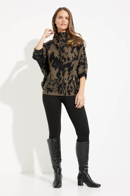 A woman stands against a plain white background, wearing an Animal Print High Collar Top Style 233948 by Joseph Ribkoff, paired with a green and black tie-dye patterned sweater, black leggings, and knee-high black boots. She has long, wavy brown hair and holds her left hand near her neck—an effortless addition to any fall wardrobe.