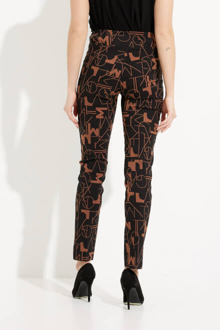 A person wearing the Abstract Print Straight Leg Pants Style 233279 by Joseph Ribkoff, featuring a black high-waisted design with an abstract brown geometric pattern, paired with a black top and black high heels, stands against a white background with one hand on their hip.