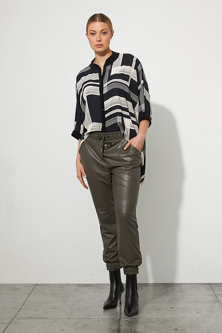 A woman stands against a plain white background, wearing a black and white geometric patterned top with three-quarter sleeves. She pairs it with Joseph Ribkoff's Faux Leather Joggers Style 223166 in Avocado and black ankle boots, with her hair pulled back.