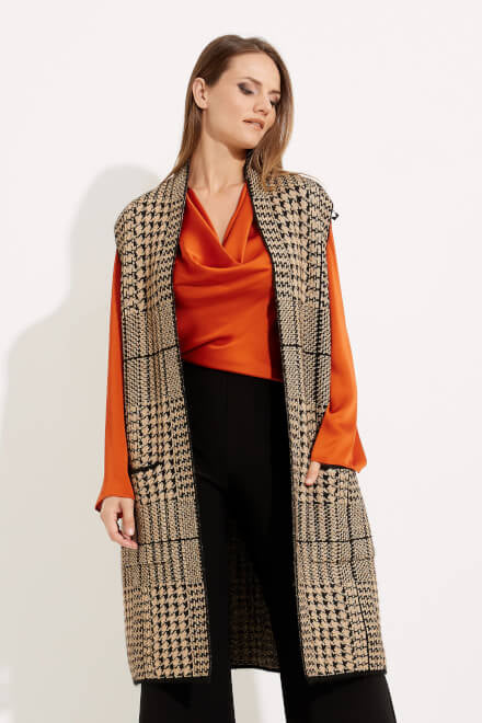 A modern woman stands against a plain white background, wearing the Long-length Houndstooth Vest Style 233957 in camel and black by Joseph Ribkoff over a silky orange blouse. She has long brown hair and is also wearing black wide-leg pants. She gazes slightly downward with a neutral expression, embodying an elegant, luxurious style.