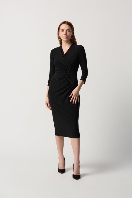 A woman with long hair, wearing a Pencil Sheath Dress Style 234272 by Joseph Ribkoff—black, knee-length, wrap-style with three-quarter sleeves and a V-neck—stands against a light grey background. She is also wearing black heels. Her left hand rests on her hip, and she looks confidently at the camera.